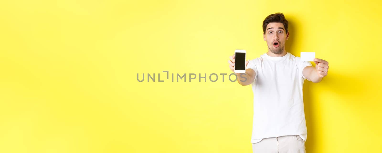 Handsome caucasian guy showing smartphone screen and credit card, concept of mobile banking and online shopping, yellow background.