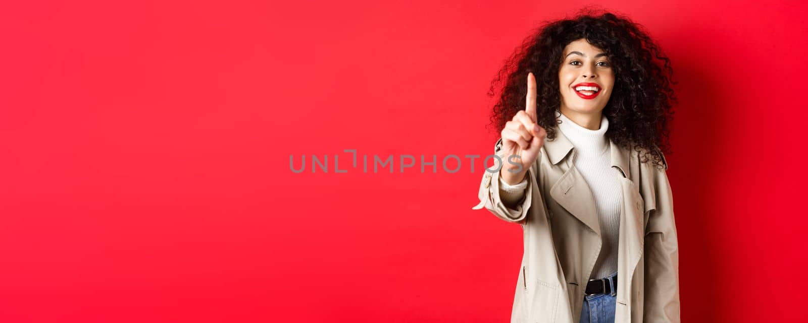 Cheerful woman in trench coat, showing number one finger and smiling, standing on red background.