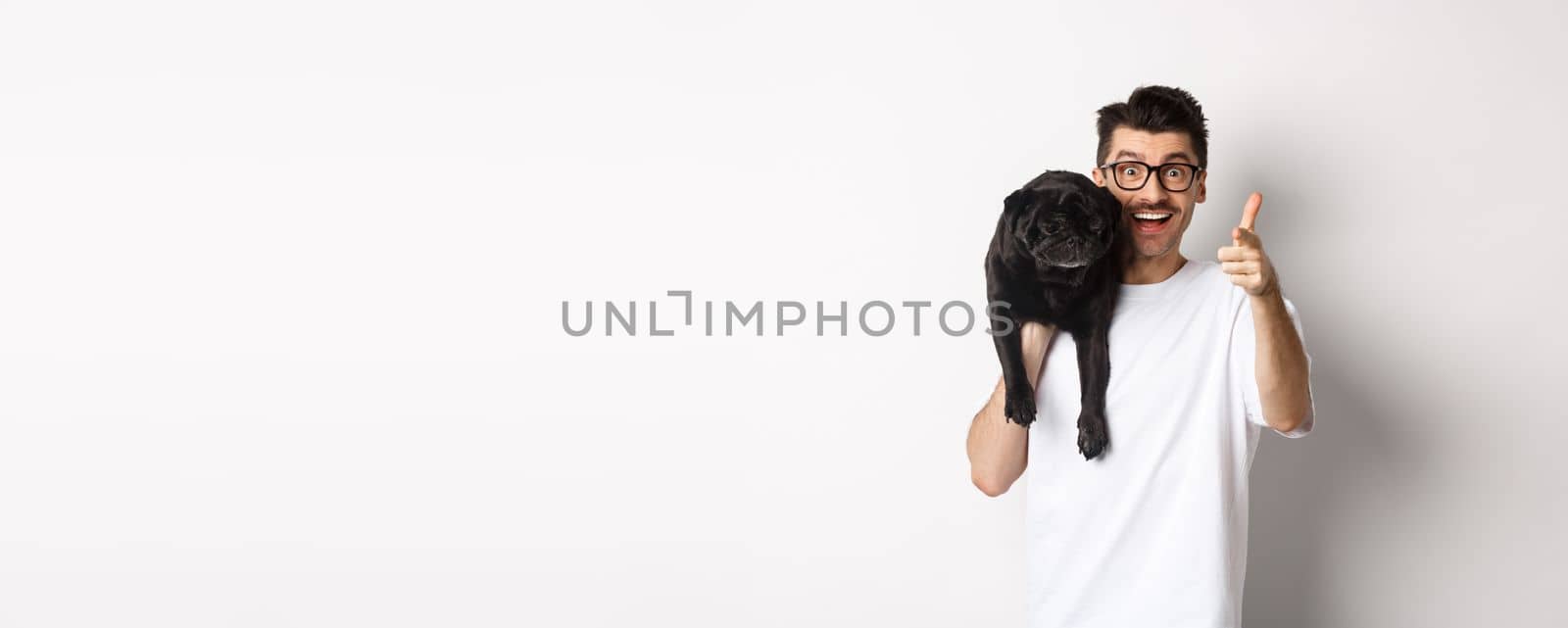 Happy young man holding cute black dog on shoulder and pointing at camera. Hipster guy carry pug on shoulder and staring at camera excited, standing over white background.