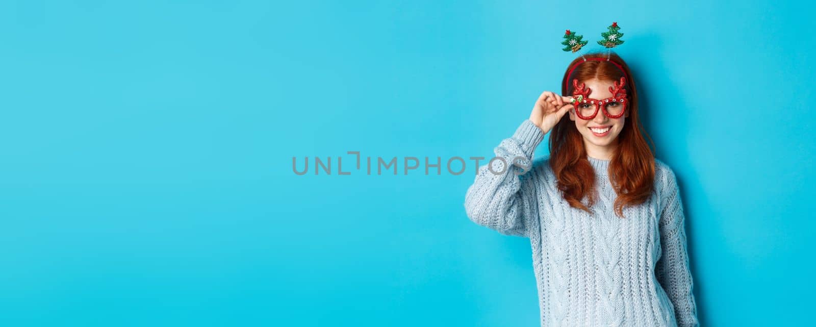 Winter holidays and Christmas sales concept. Beautiful redhead female model celebrating New Year, wearing funny party headband and glasses, smiling at camera by Benzoix