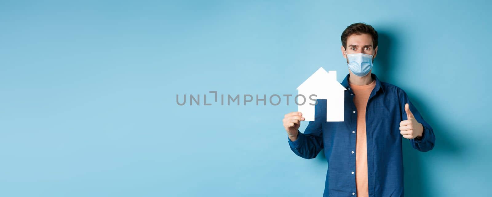 Real estate and covid concept. Young man in face mask showing paper house cutout and thumb-up, recommending agency, standing on blue background.