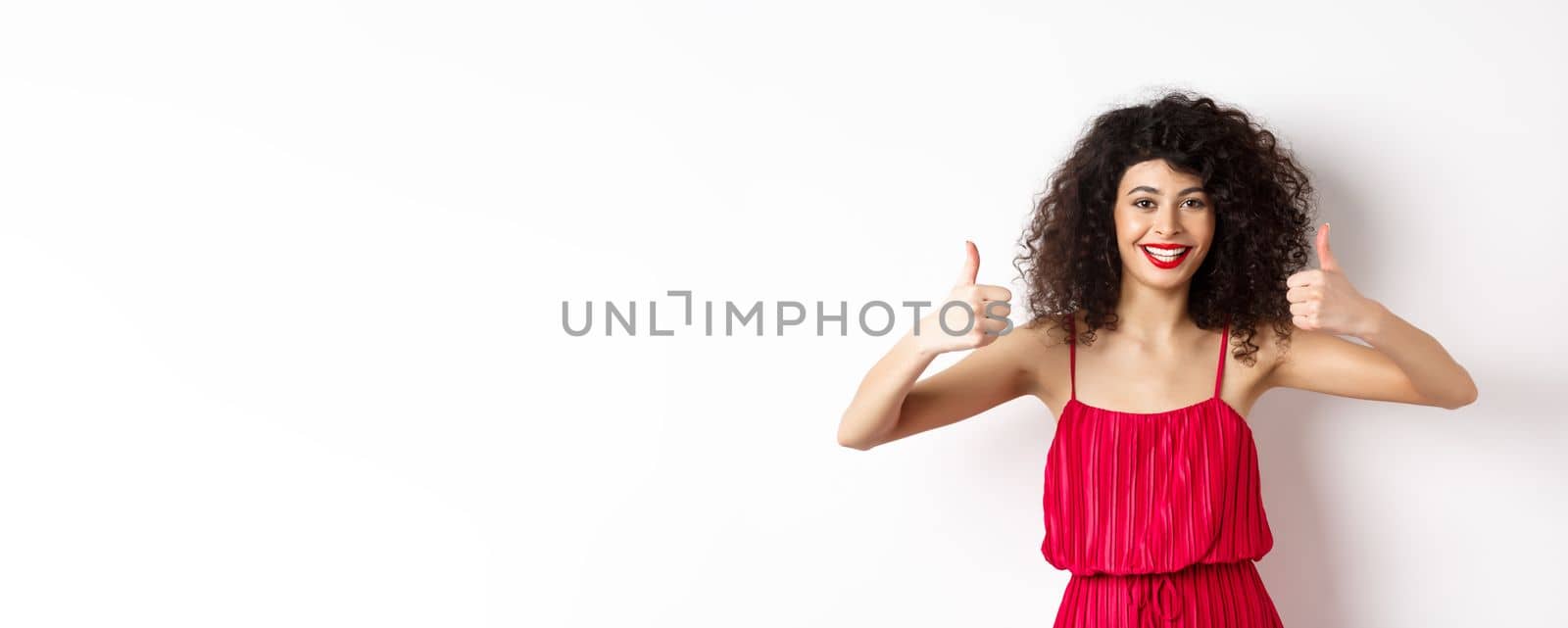 Attractive young woman recommending promo offer, showing thumb up and smiling, like product, standing in festive red dress on white background by Benzoix