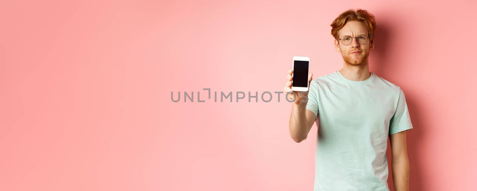 Disappointed male model frowning, showing smartphone screen, standing over pink background by Benzoix