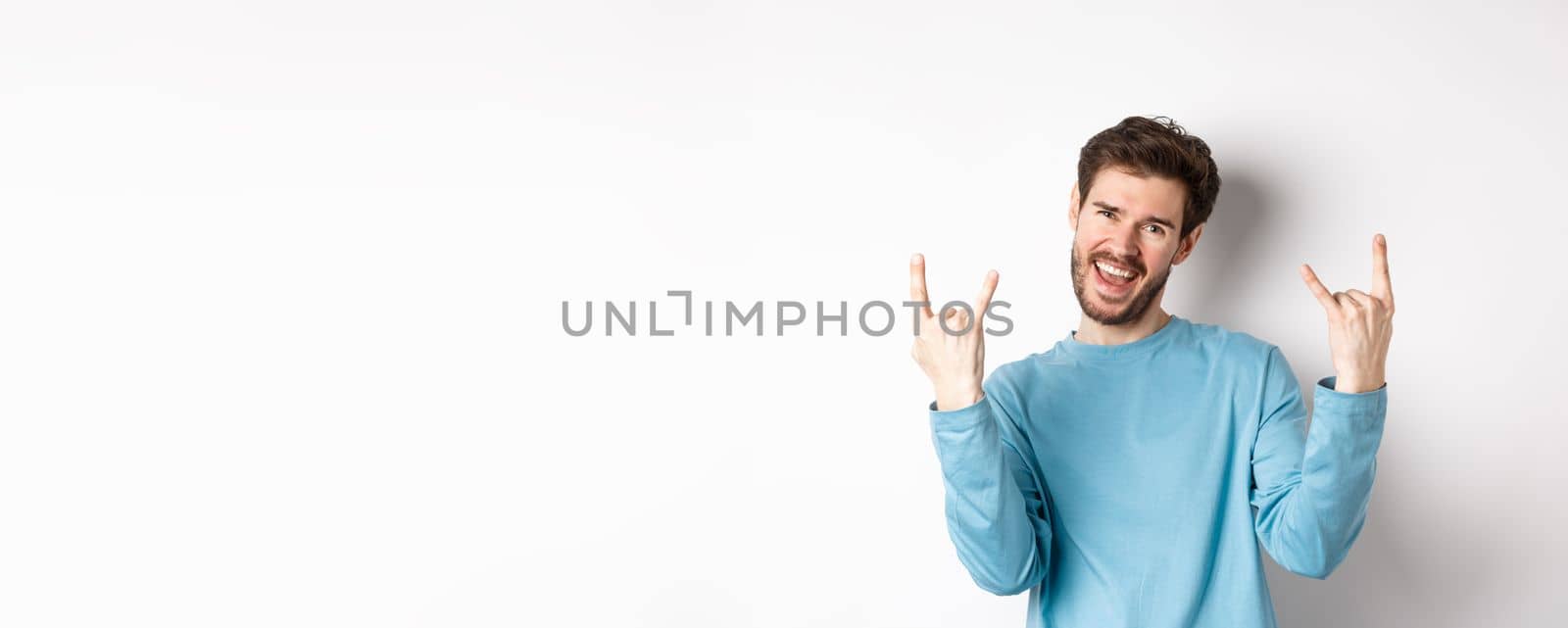 Excited handsome man celebrating, having fun and showing rock on horns gesture, enjoing party, smiling at camera, standing on white background.