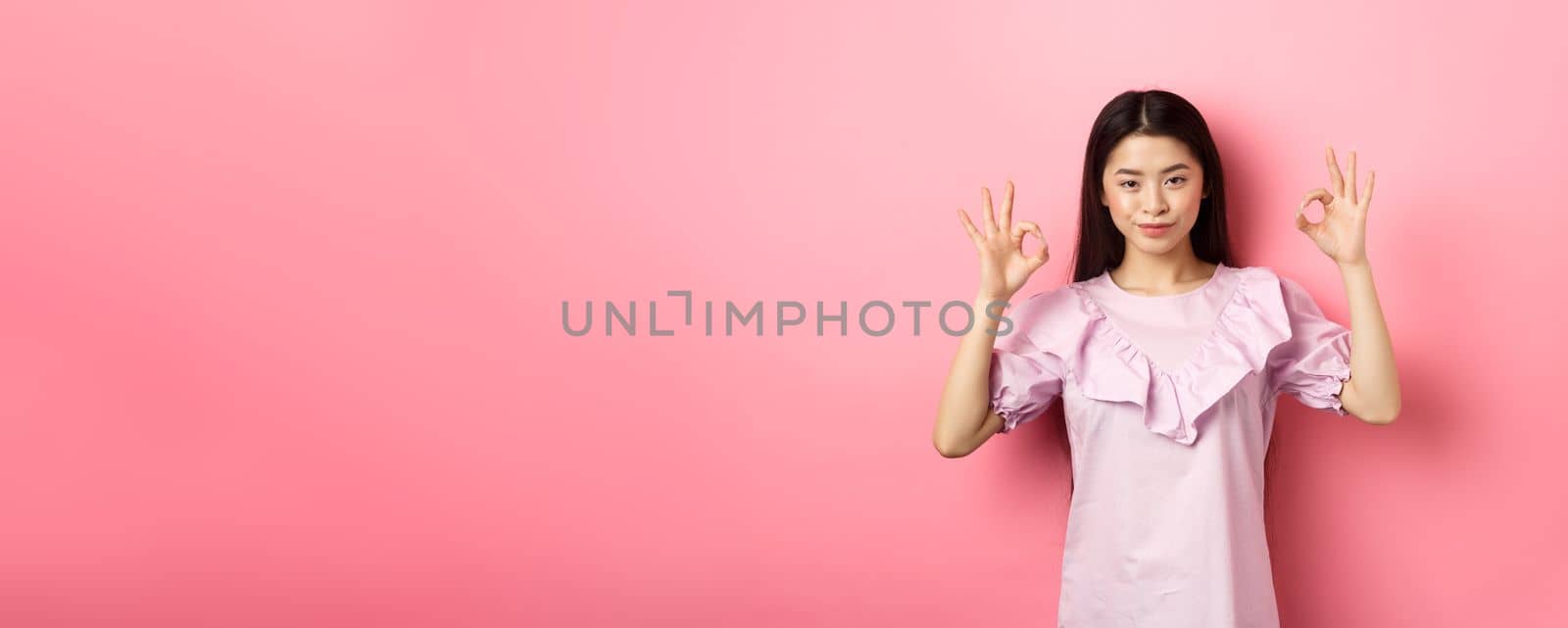 Smiling asian woman showing okay signs and looking confident, assure all good, praise good work, nice choice gesture, standing on pink background.