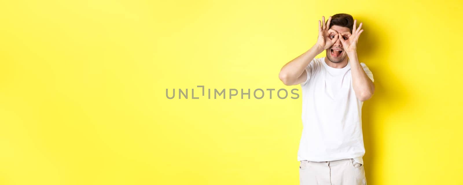 Young man showing funny faces and sticking tongue, standing playful against yellow background.