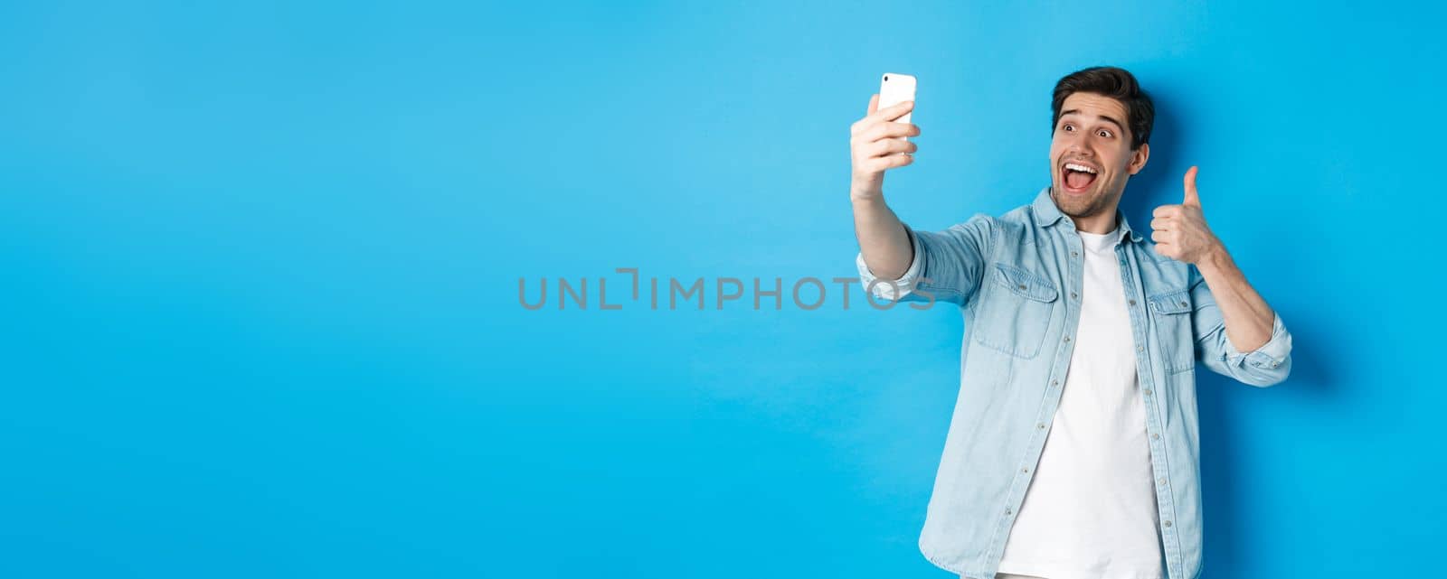 Happy man taking selfie and showing thumb up in approval on blue background, holding mobile phone.