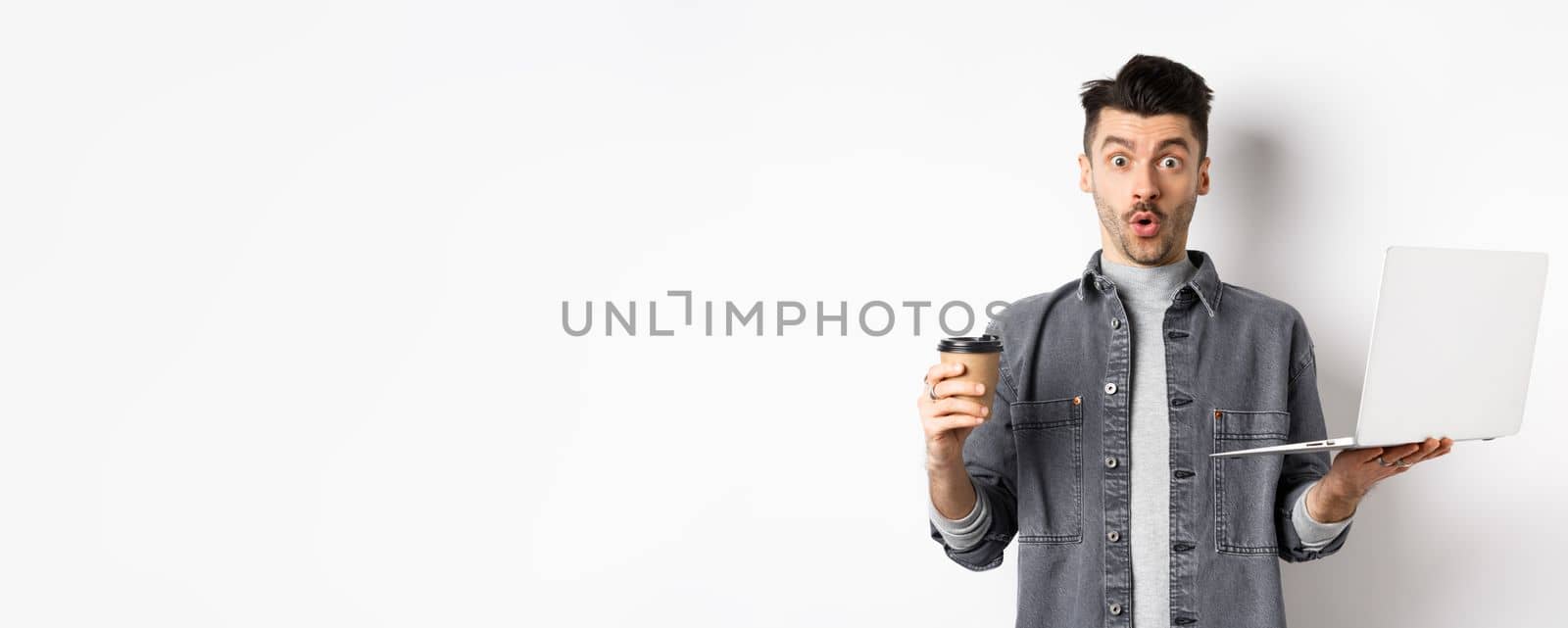 Excited man say wow, holding laptop for work and coffee in paper cup, standing on white background.