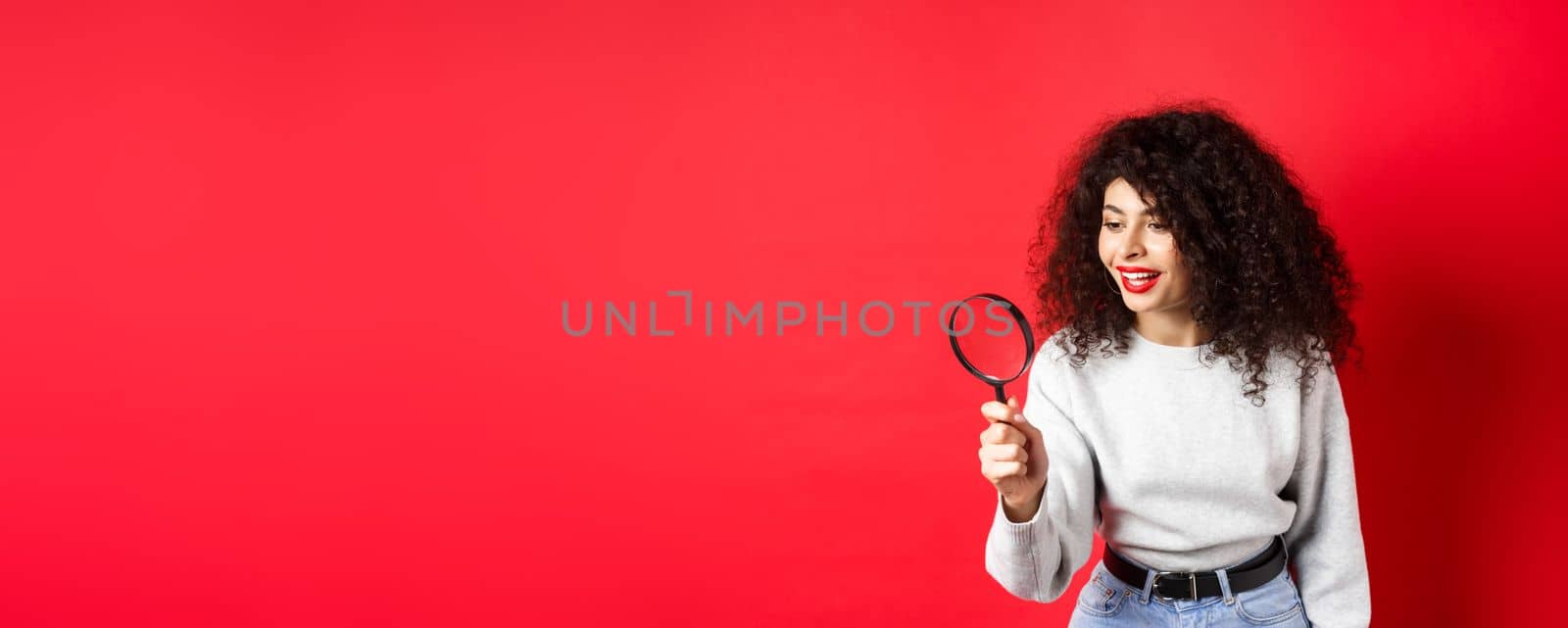 Young woman looking through magnifying glass, investigating or searching for something, stare curious aside, found interesting thing, standing on red background by Benzoix