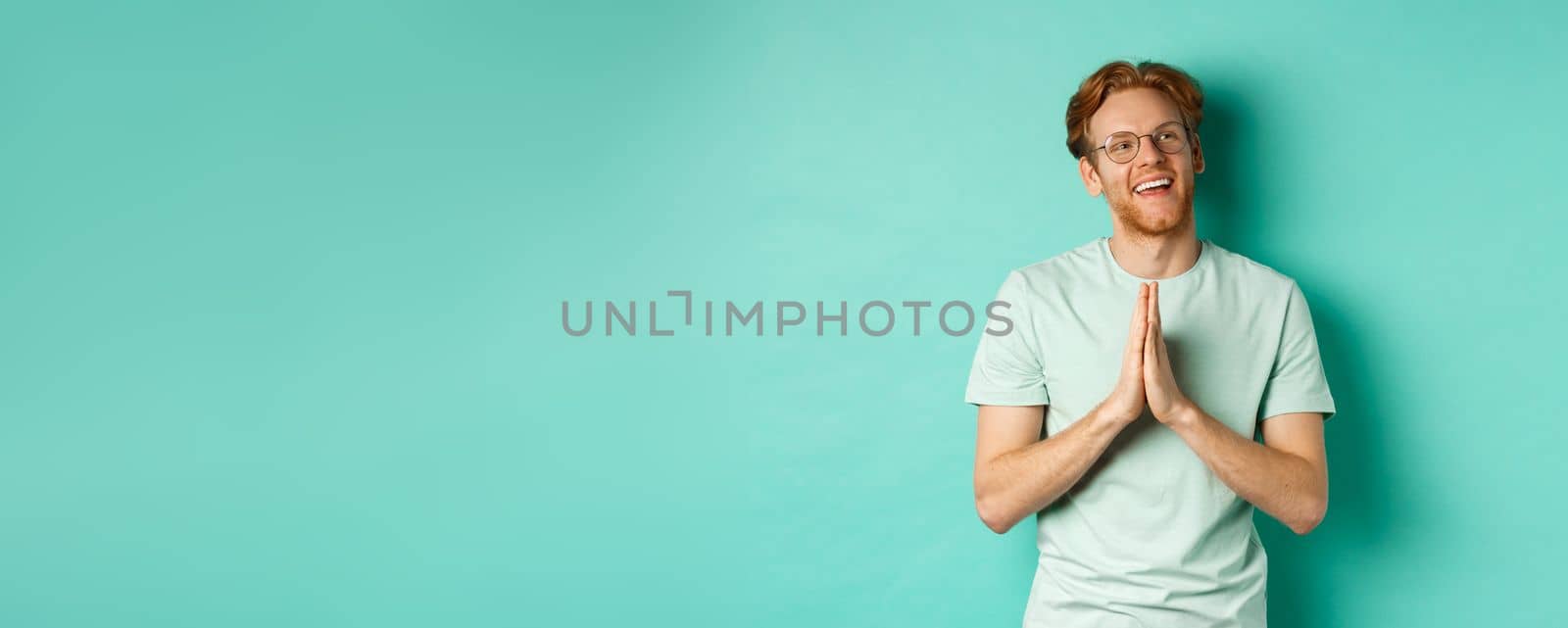 Hopeful redhead man with beard, wearing glasses and t-shirt, holding hands in namaste or plead gesture and looking right, smiling and thanking, standing over turquoise background.