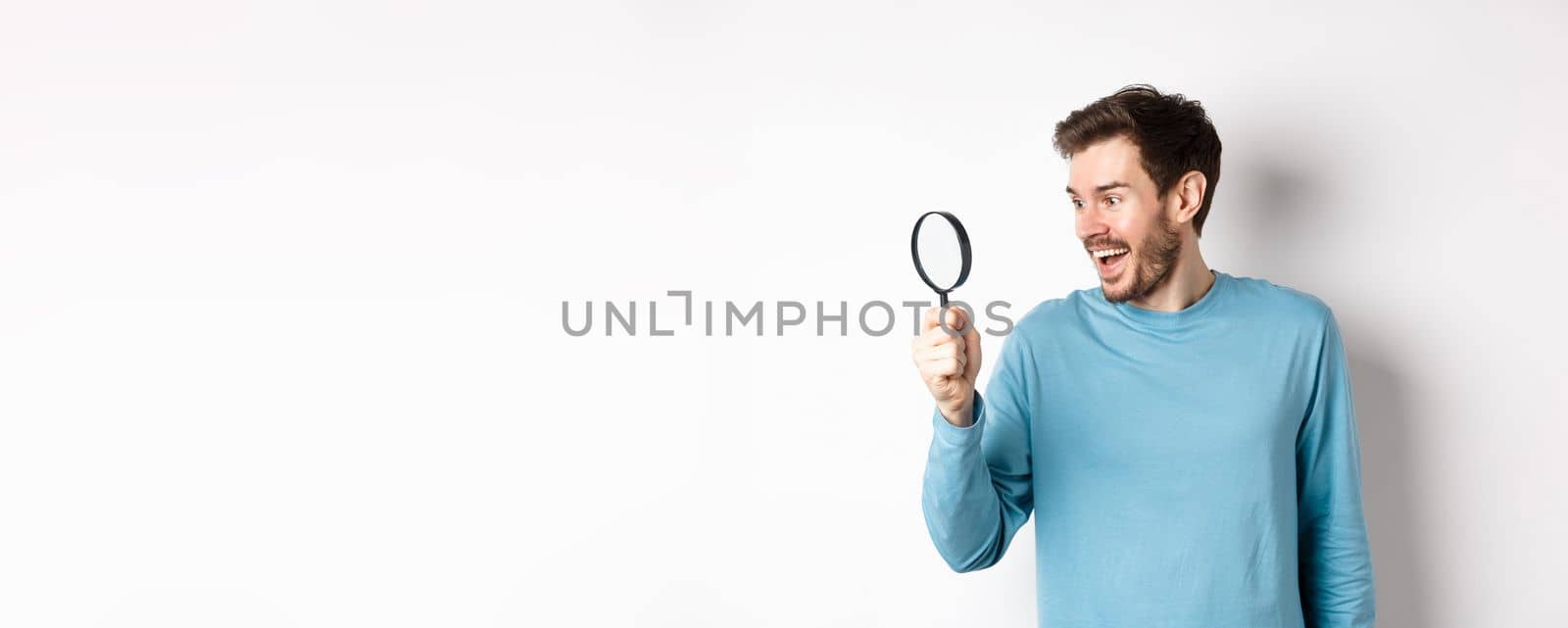 Handsome smiling man looking through magnifying glass with amazed face, found interesting promotion, standing on white background.
