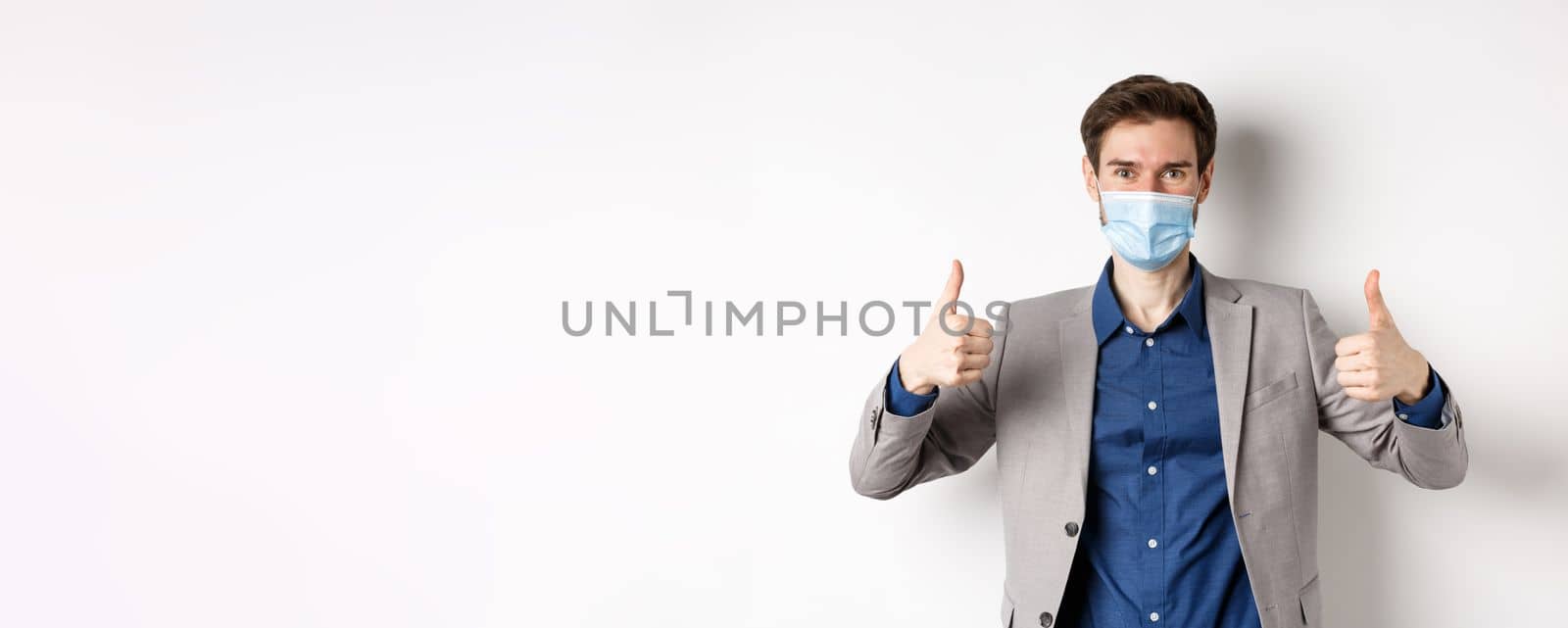 Covid-19, pandemic and business concept. Cheerful man in suit and medical mask using preventive measures in office, showing thumbs up, white background.