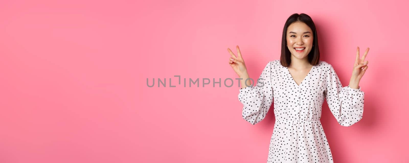 Cute asian brunette girl in dress smiling, showing kawaii peace signs and looking happy, standing over pink background.