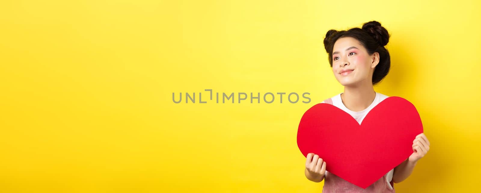 Asian girl in love, looking dreamy left and smiling, showing Valentines red heart cutout, imaging romantic date, standing on yellow background.