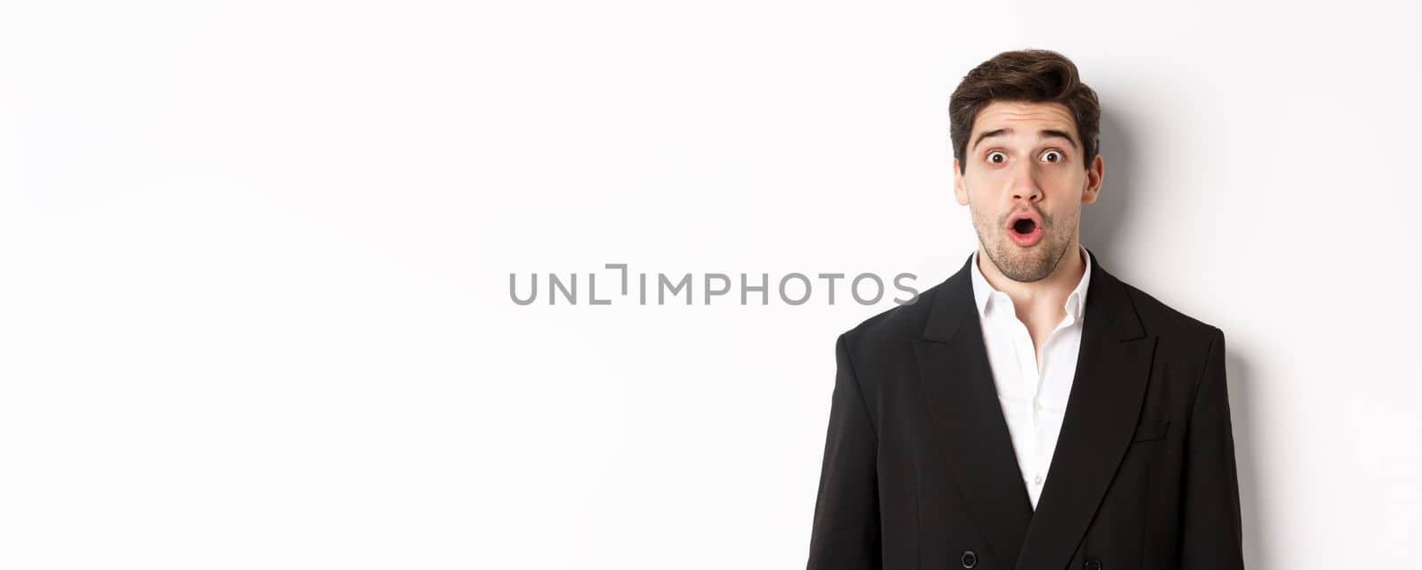 Close-up of attractive man in black suit, looking surprised and impressed at advertisement, standing over white background.