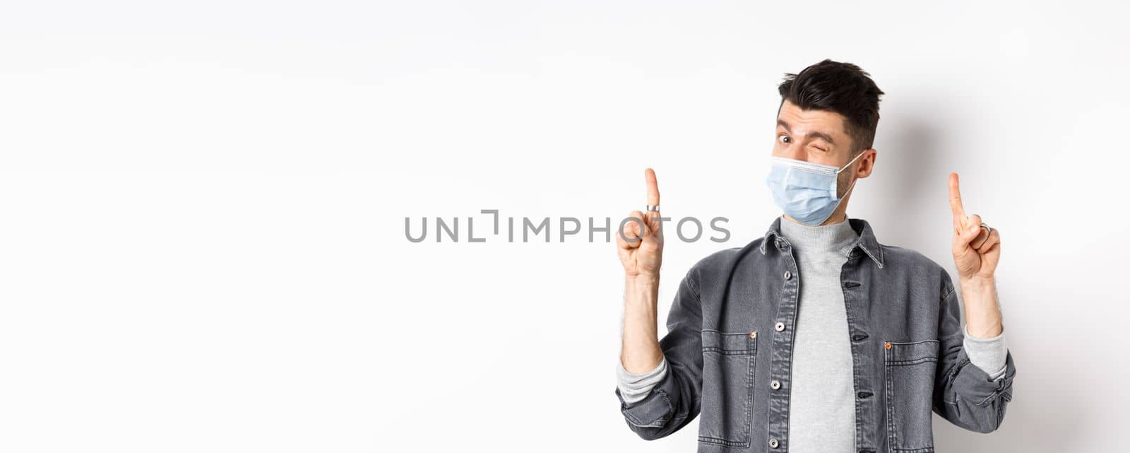 Pandemic lifestyle, healthcare and medicine concept. Handsome young man winking in medical mask, make announcement, showing promo deal, pointing fingers up, white background.
