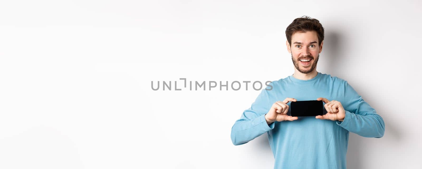 E-commerce and shopping concept. Smiling cheerful man showing empty smartphone screen in horizontal position, looking happy at camera, white background.