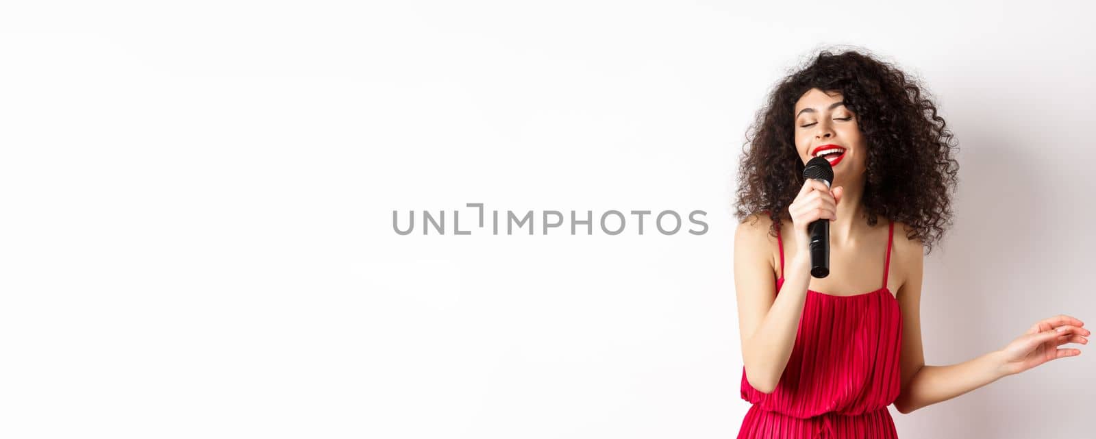 Happy elegant woman in red dress performing with microphone, singing karaoke and smiling, standing over white background by Benzoix