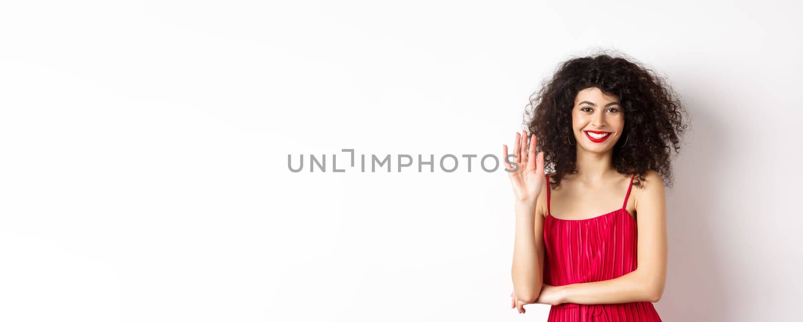 Cheerful elegant woman saying hello, waving hand and smiling at camera, greeting someone, standing in red dress on white background.