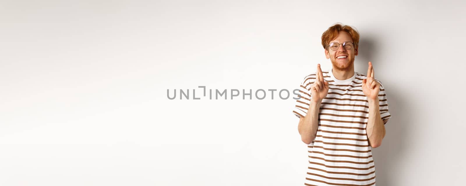 Happy redhead man with bristle cross fingers for good luck and smiling hopeful, making wish, standing over white background.