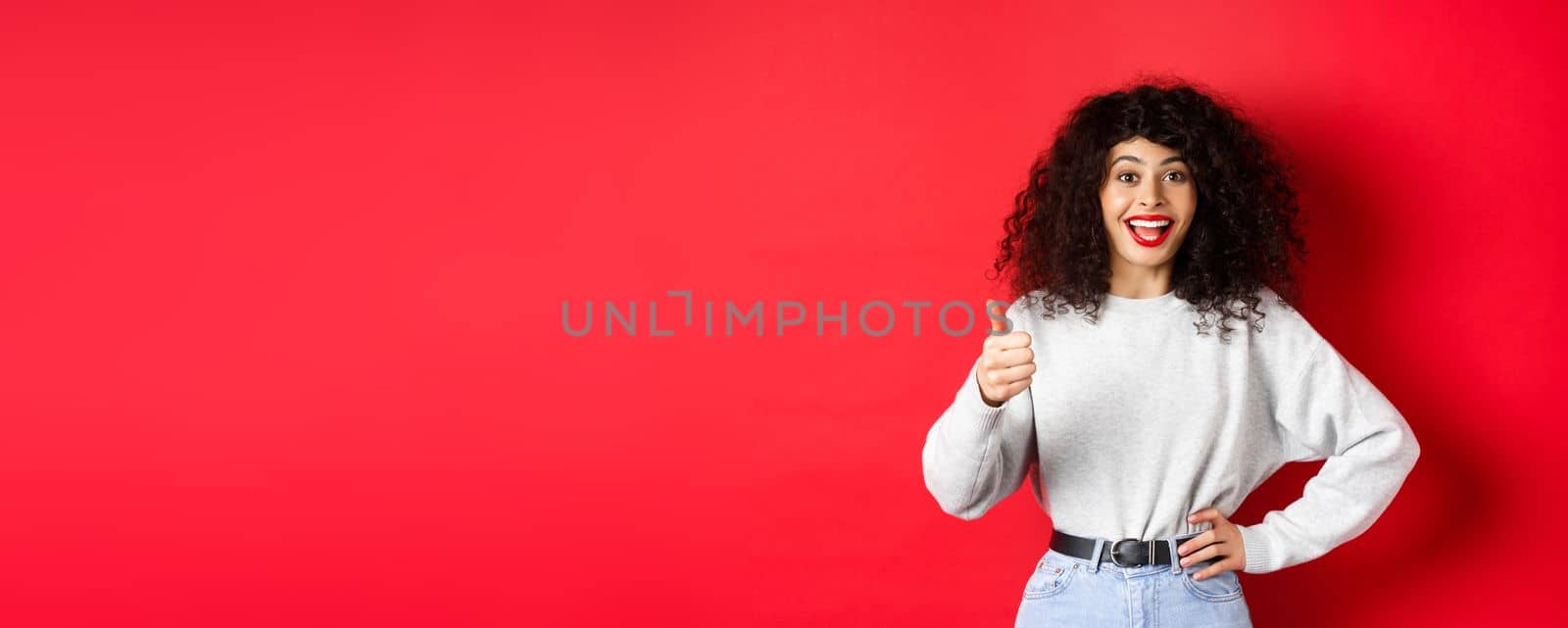 Happy young woman with curly hair praising good work, say well done and show thumb up gesture, approve and praise you, standing on red background by Benzoix