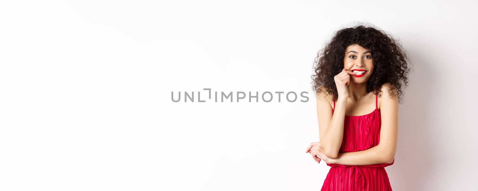 Intrigued young woman with curly hair, wearing elegant red dress and lipstick, biting fingernails and looking interested, standing over white background.