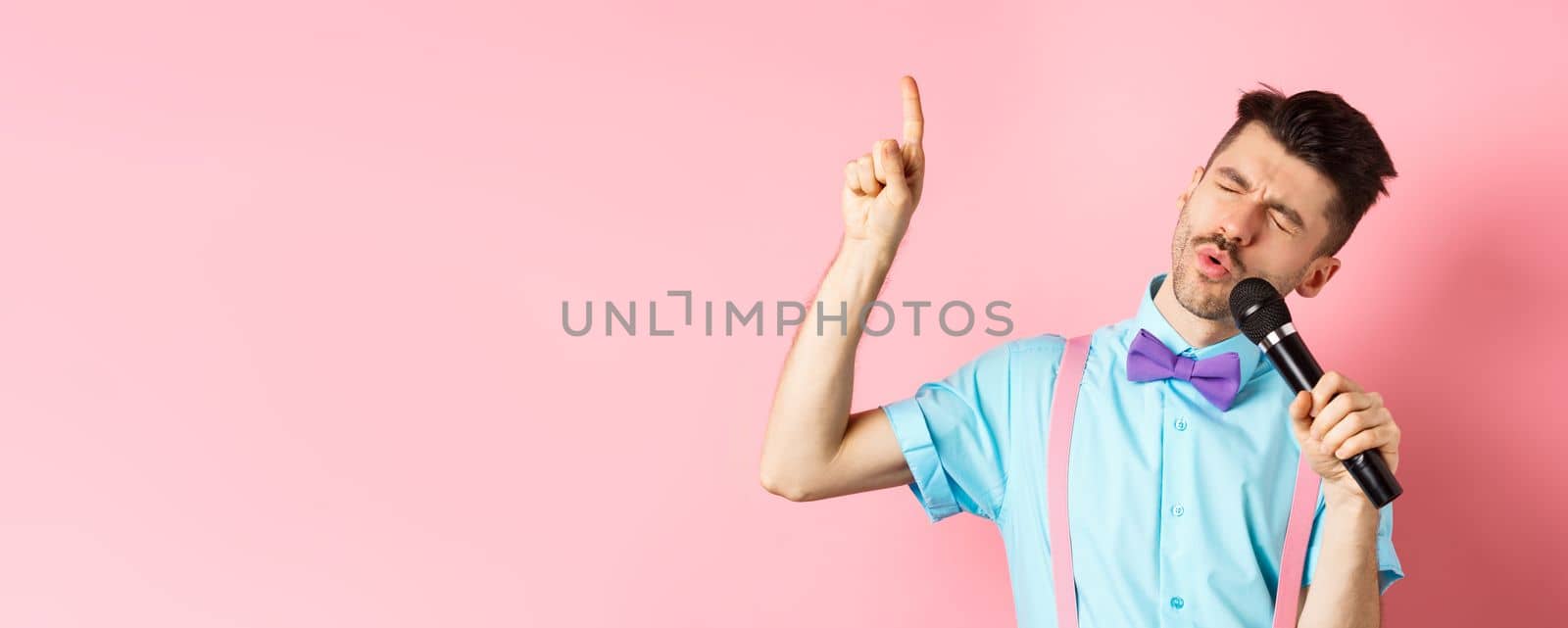 Party and festive events concept. Funny guy singing in microphone, raising finger up as reaching high note in song, standing on pink background.
