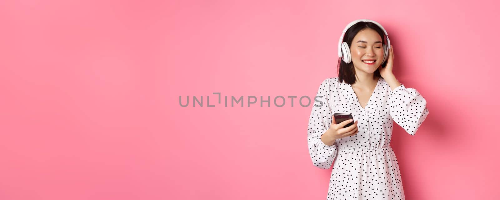 Romantic asian girl listening music in headphones, smiling with closed eyes, holding mobile phone, standing over pink background.