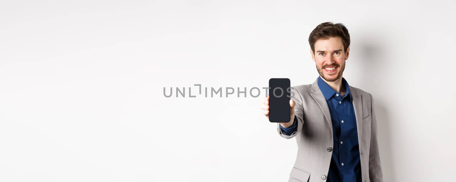E-commerce and online shopping concept. Confident businessman in suit stretch out hand with empty smartphone screen, showing on phone, standing against white background.