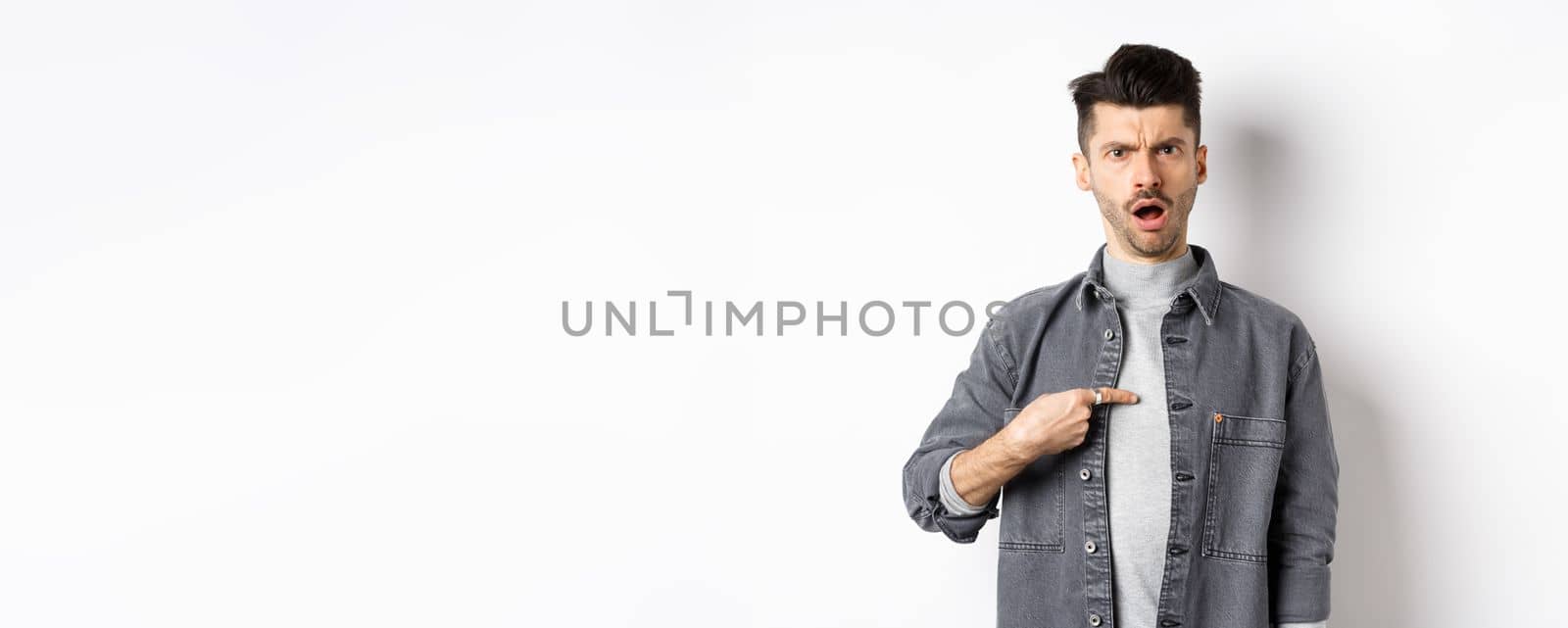 Offended young man being accused, frowning and pointing at himself shocked, standing on white background.