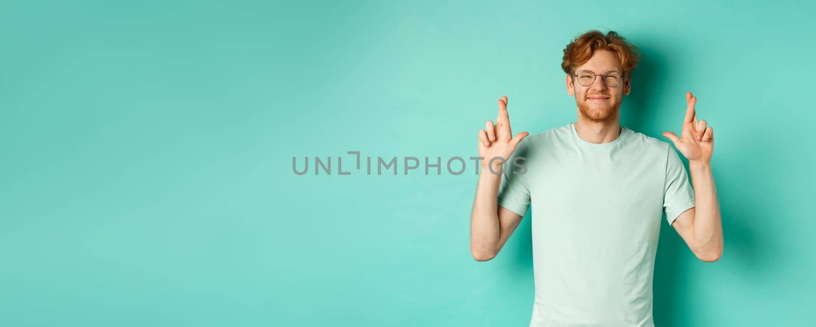 Young cheerful guy with red hair and beard, wearing glasses, smiling and cross fingers for good luck, making wish and looking optimistic, standing over mint background by Benzoix