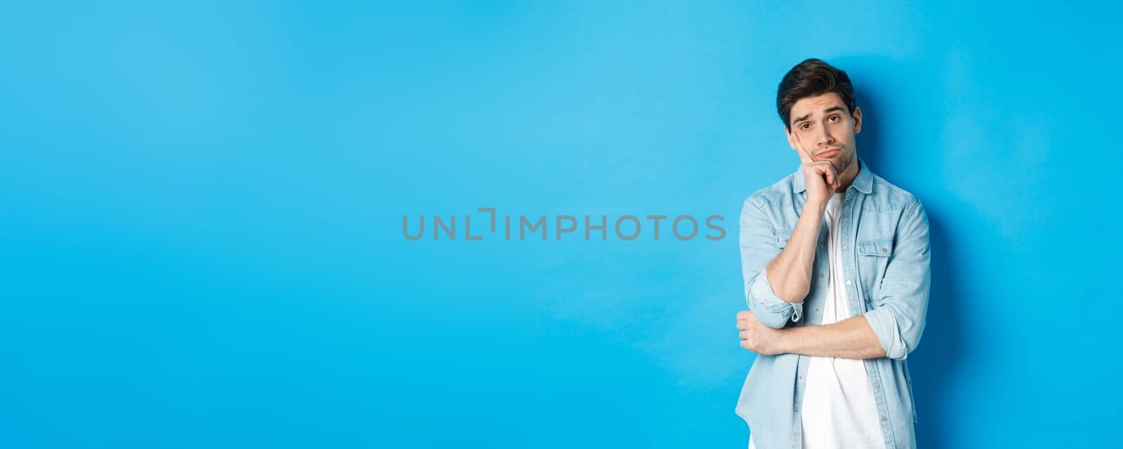 Unamused and bored man looking without interest at camera, standing against bllue background by Benzoix