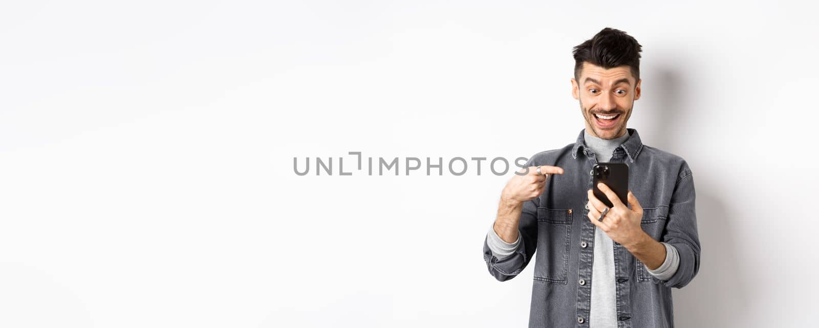 Excited handsome man showing cool news on phone, pointing at smartphone and smiling happy, standing against white background.