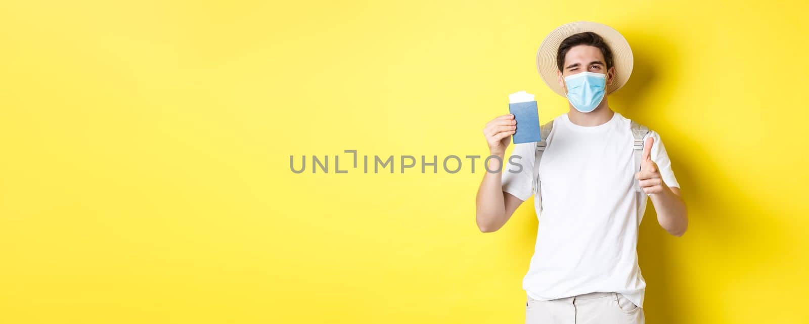 Concept of covid-19, tourism and pandemic. Happy guy tourist in medical mask showing passport with tickets for vacation, winking and pointing at camera, yellow background.