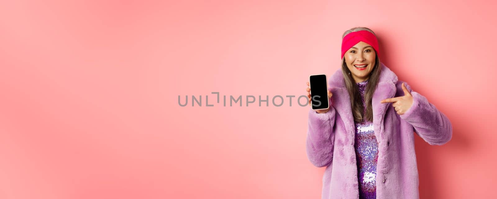 Online shopping and fashion concept. Stylish asian mature lady showing blank smartphone screen, pointing at phone and smiling, pink background.