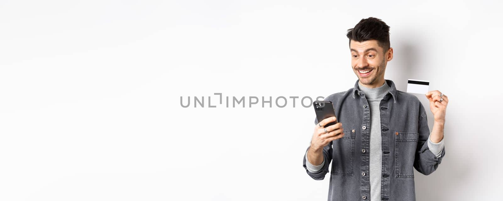 Online shopping concept. Smiling guy paying on mobile phone, showing plastic credit card, buying in internet, white background by Benzoix