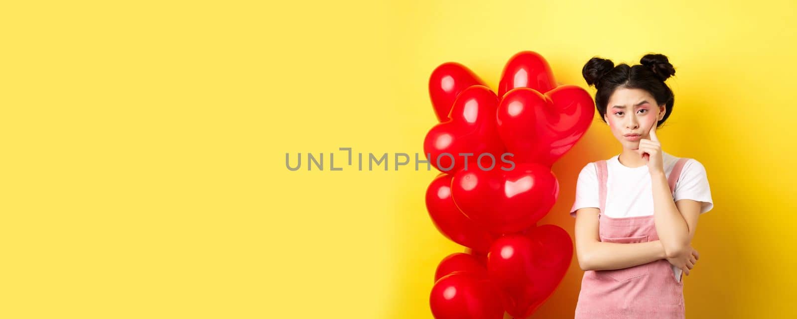 Valentines day concept. Unamused and bored asian girlfriend staring skeptical at camera, waiting for something near red hearts balloons, yellow background.