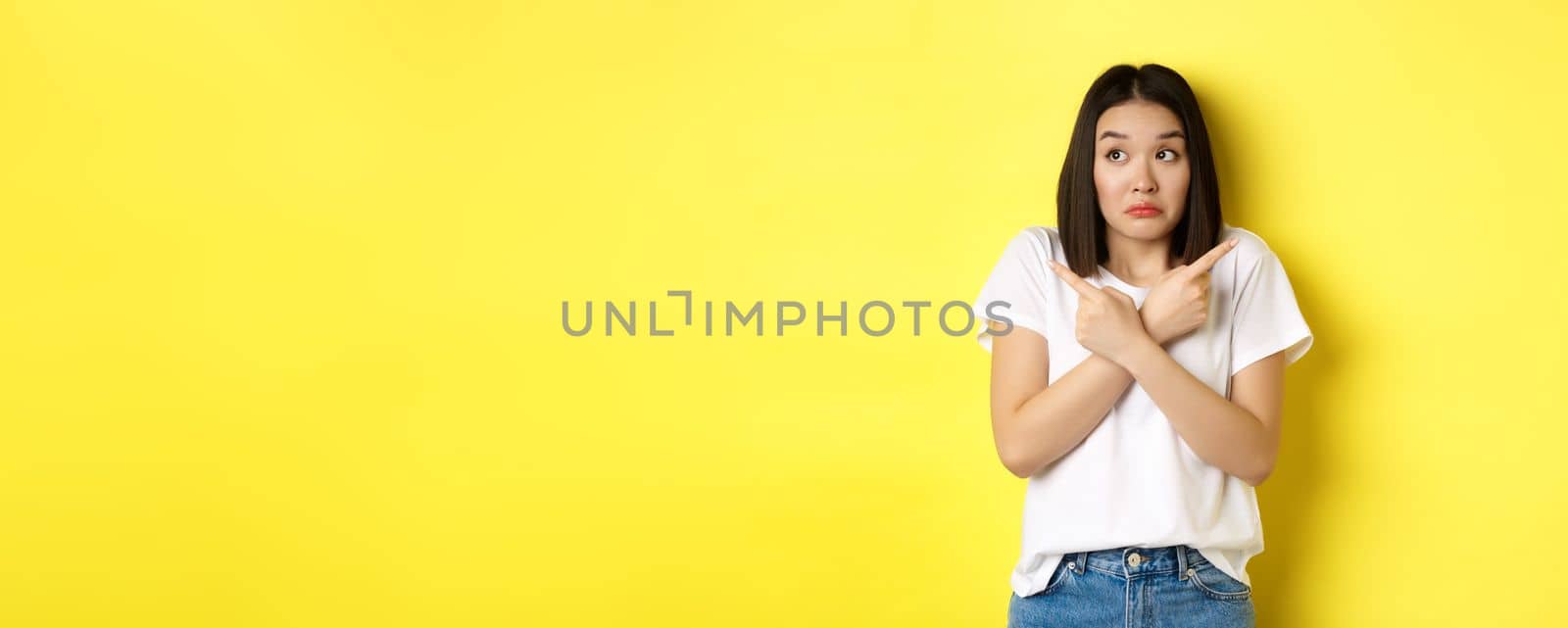 Indecisive asian girl need help with choice, pointing fingers sideways and looking confused, standing over yellow background.