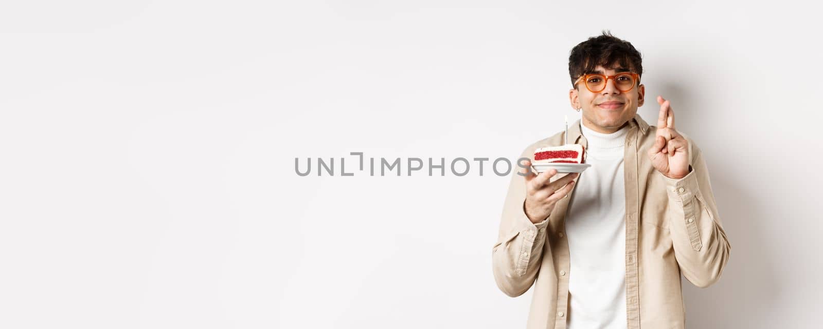 Wishful handsome guy in glasses making wish on birthday cake, standing with fingers crossed and happy smile on white background by Benzoix
