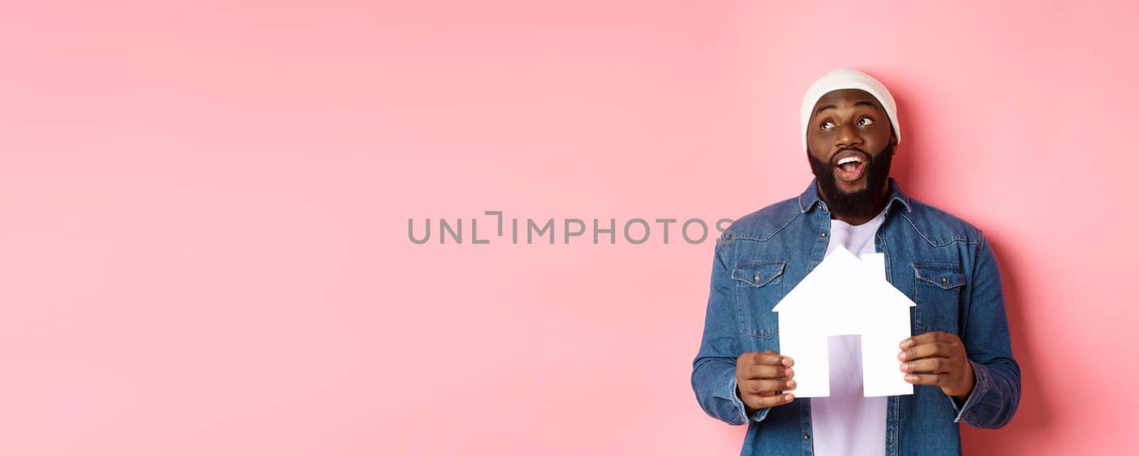 Real estate concept. Handsome african-american man dreaming about home, holding house model and looking at upper left corner, imaging apartment, pink background by Benzoix