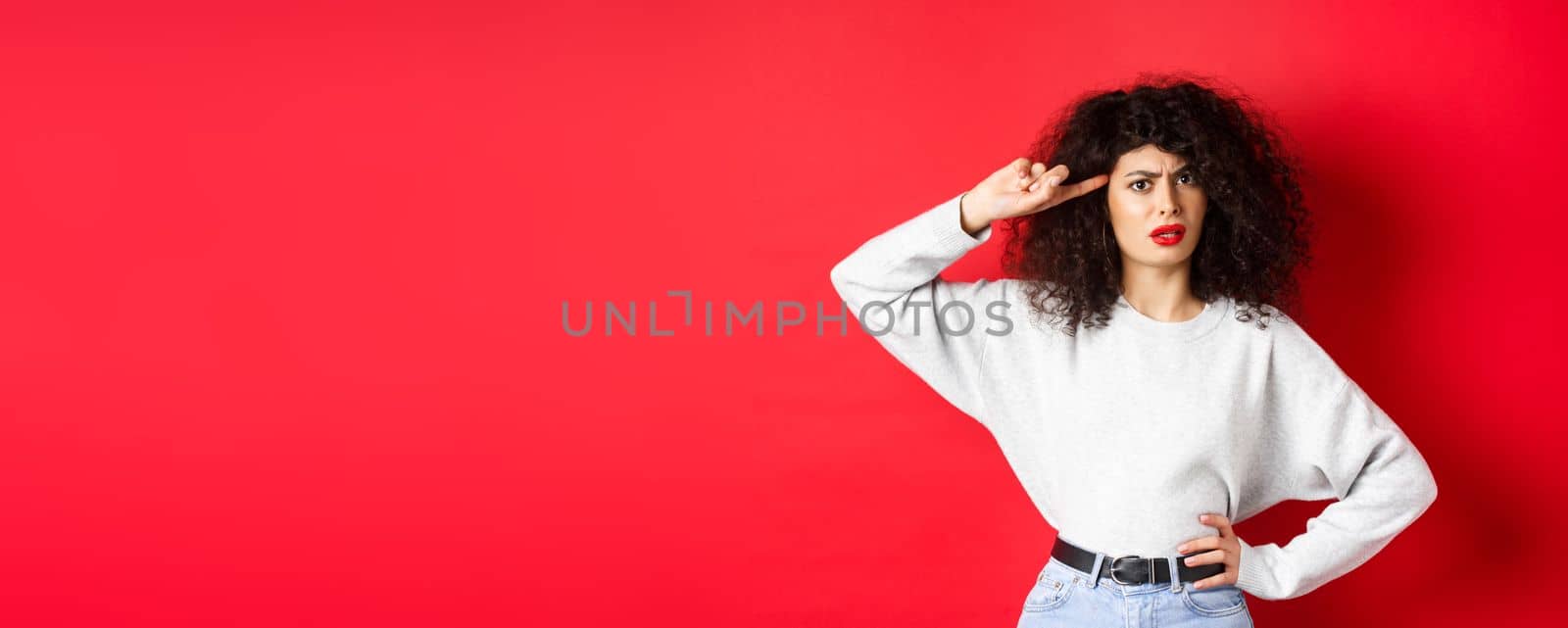 Are you stupid. Annoyed and confused girl with curly hair, pointing at head and scolding person being crazy or strange, standing on red background.