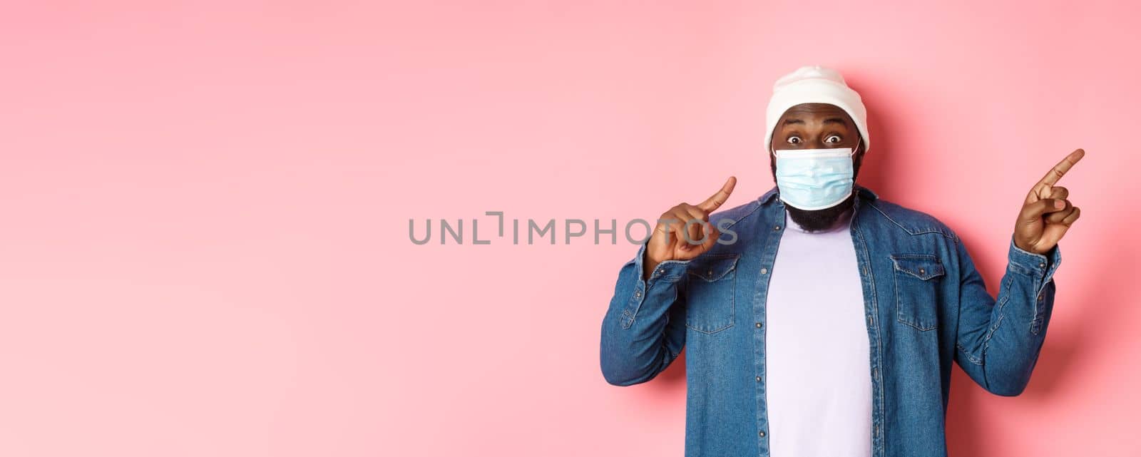 Covid-19, lifestyle and lockdown concept. Impressed Black man in face mask showing announcement, pointing right at promo and staring at camera amazed, pink background.