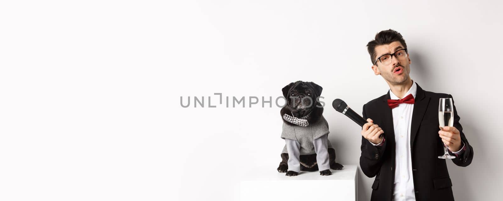 Handsome fancy man in glasses, raising glass of champagne and giving microphone to cute pug in party suit, celebrating and having fun, white background.