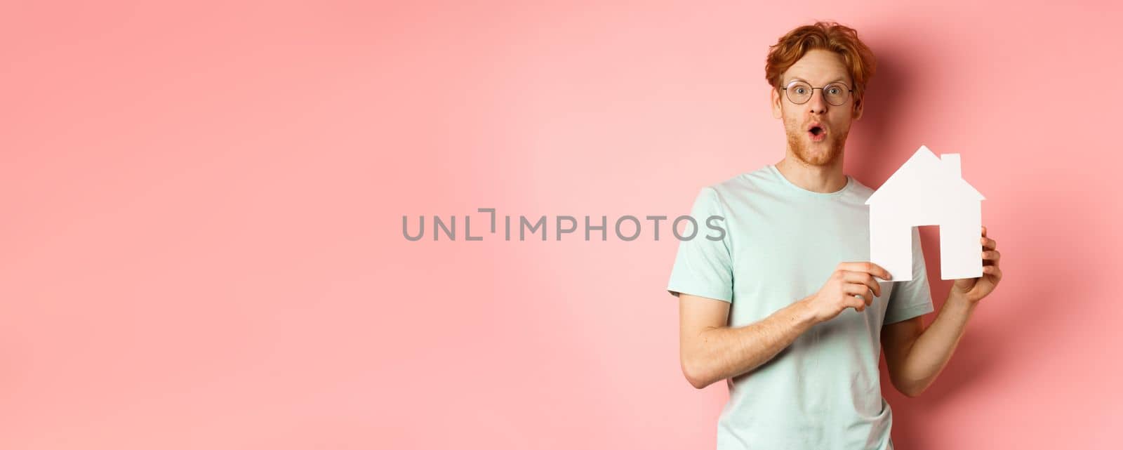 Real estate. Surprised young man with red hair and beard, wearing glasses and t-shirt, showing paper house cutout and looking impressed, saying wow, standing over pink background by Benzoix
