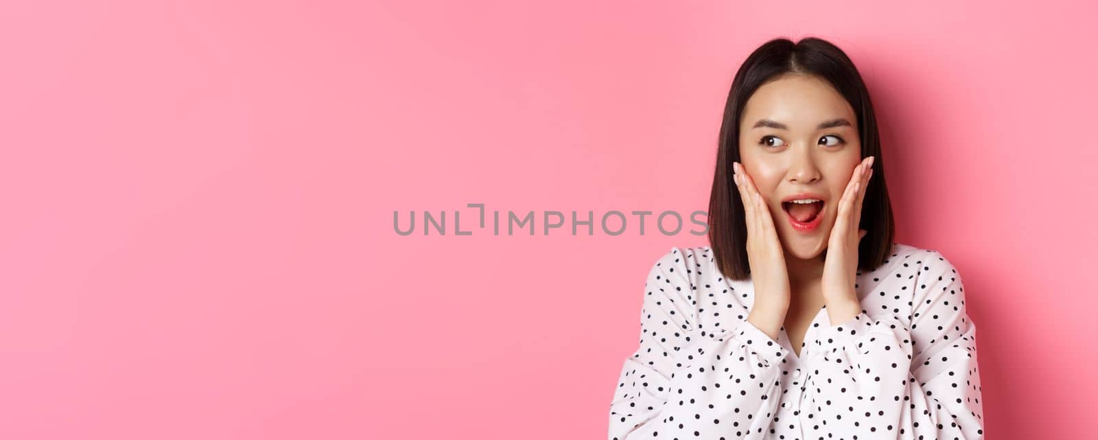 Close-up of beautiful asian woman looking surprised and excited, hear amazing news, looking left and rejoicing, standing against pink background.