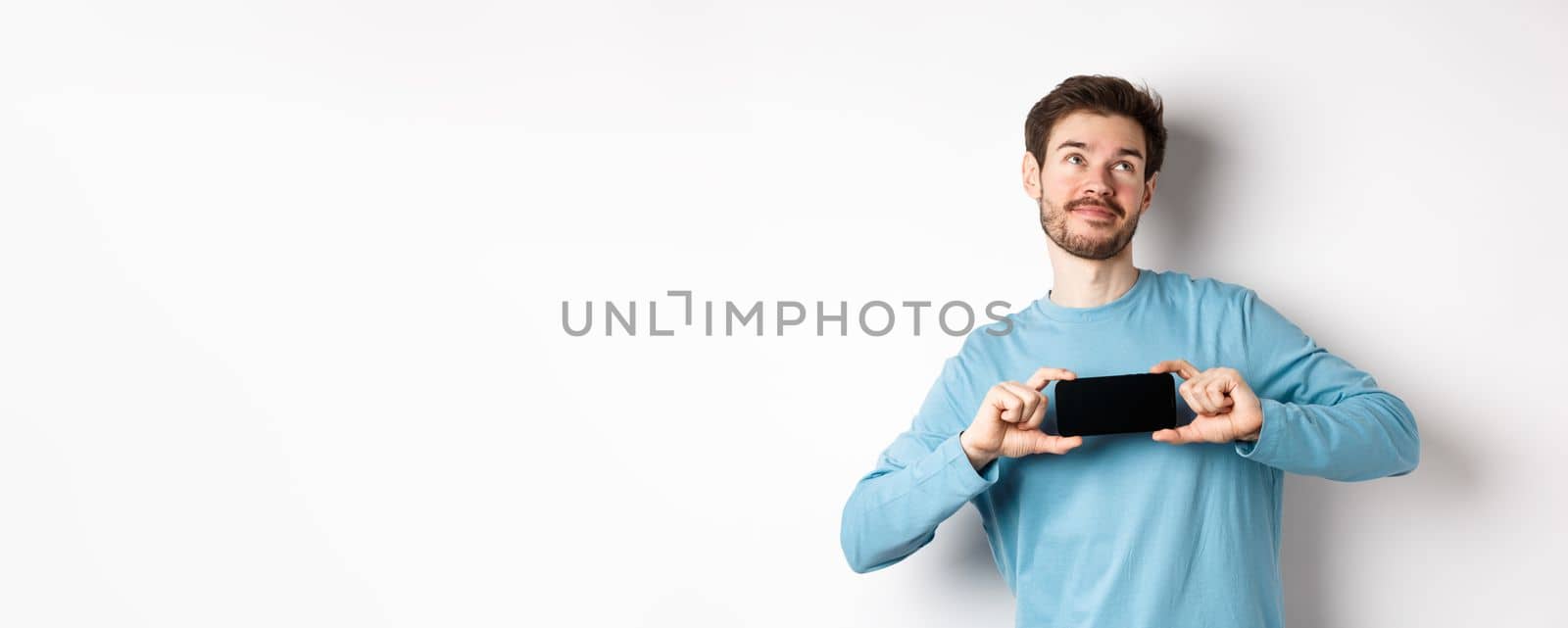 E-commerce and shopping concept. Smiling young man showing mobile screen and dreaming about something, looking at upper right corner nostalgic, standing over white background by Benzoix