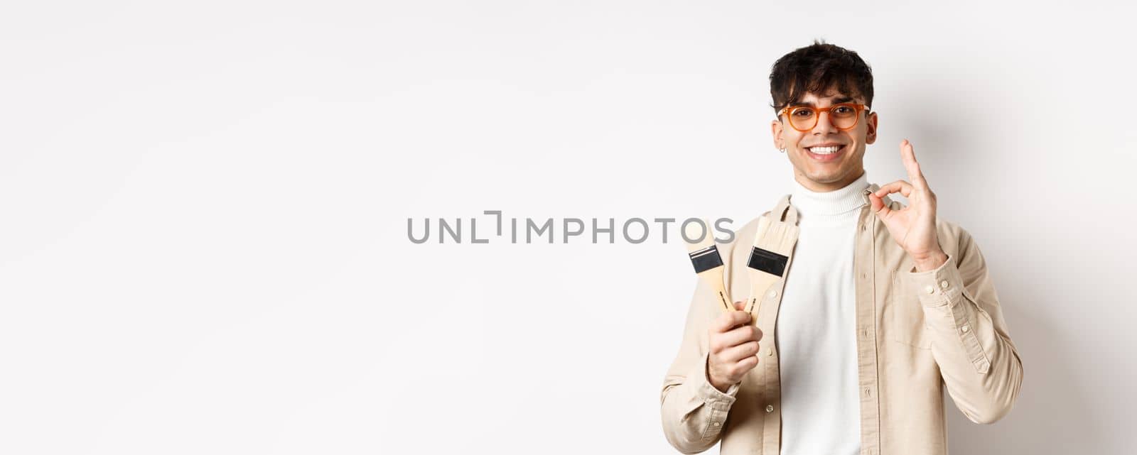 Handsome natural guy in glasses recommending store, showing painting brushes for renovation and decor, make okay sign in approval, standing on white background by Benzoix