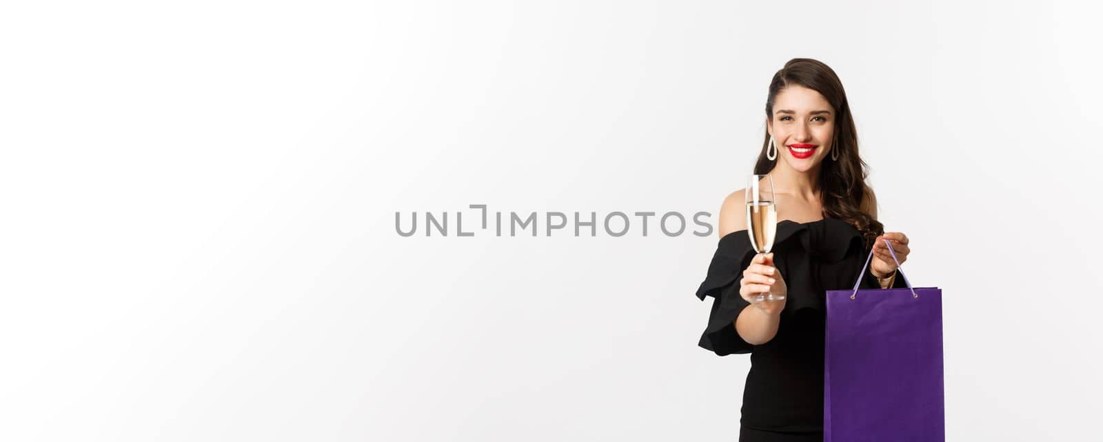 Beautiful and stylish brunette woman raising glass of champagne, celebrating christmas, holding shopping bag with presents, standing over white background.