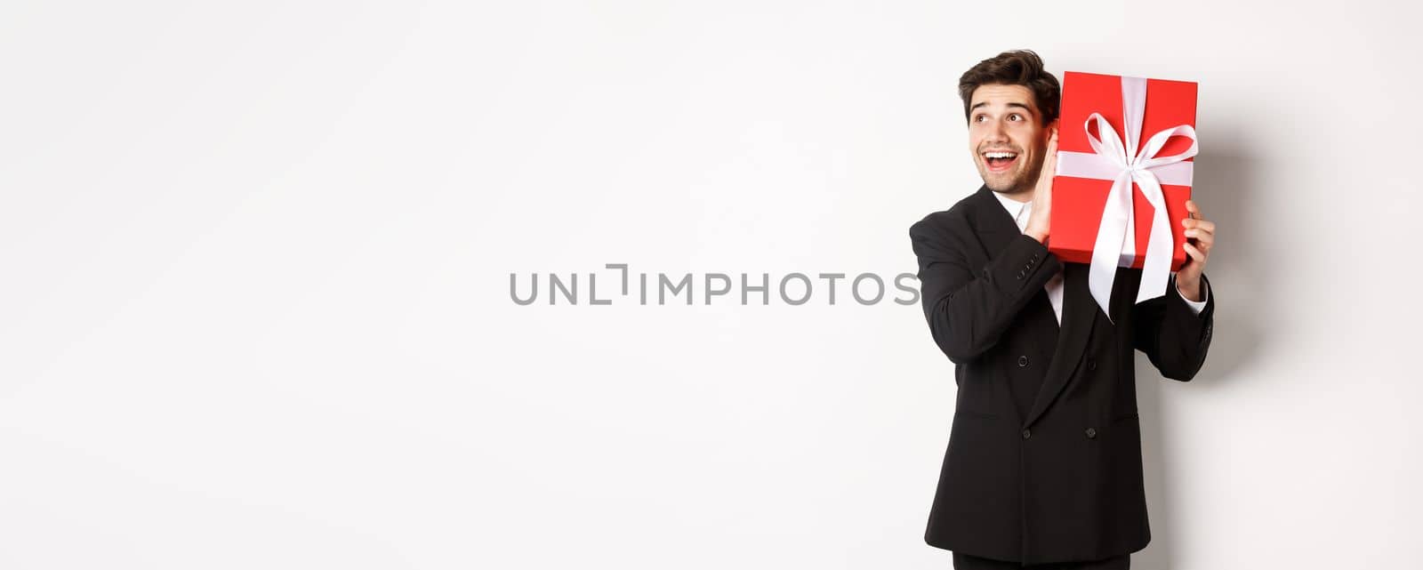 Image of handsome dreamy guy in black suit, shaking box with present to wonder whats inside, standing against white background happy.