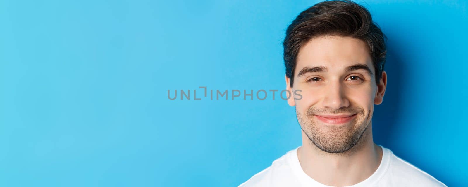 Headshot of attractive man smiling pleased, looking intrigued, standing over blue background.